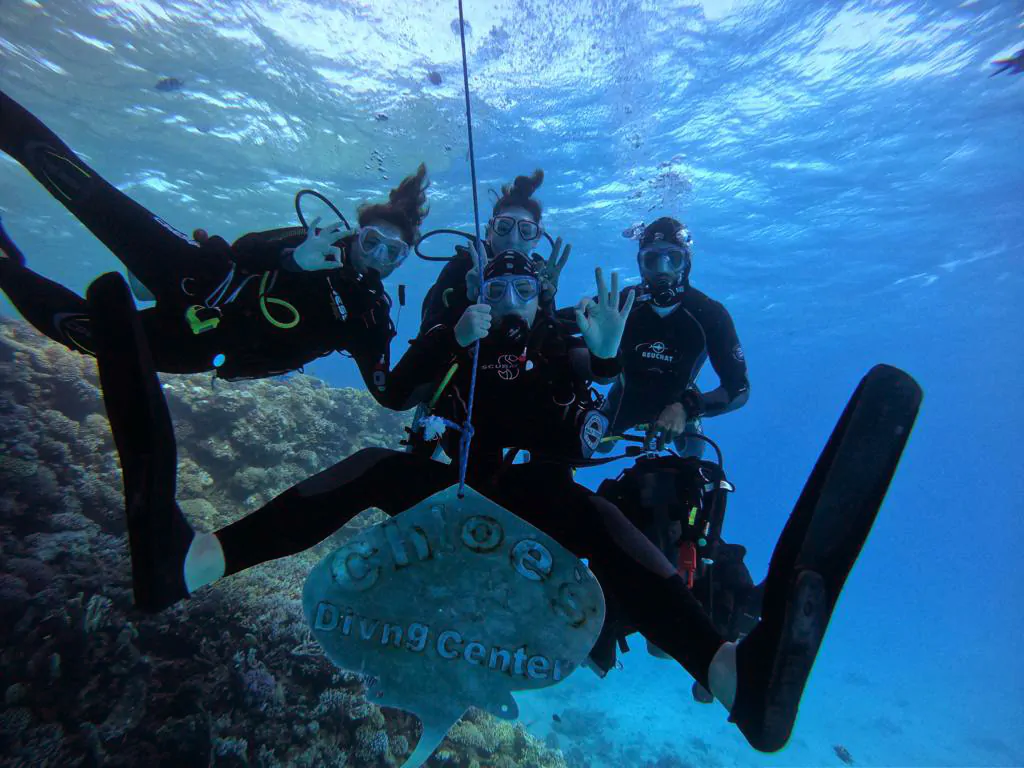 Une image de trois plongeurs et de leur guide de plongée devant le panneau du bateau du centre de plongée Chloes.